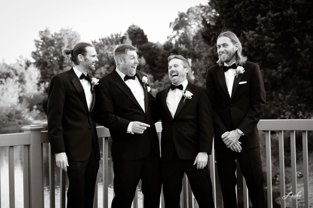 Groom and his Groomsmen stand on a deck over the water laughing on his wedding day.